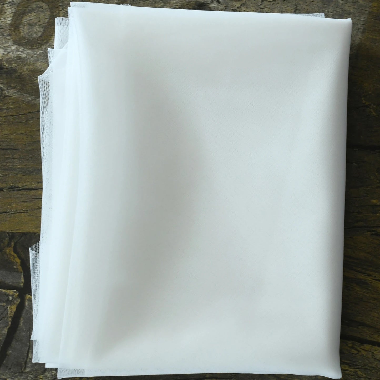 A neatly folded piece of white fabric rests on a textured wooden surface, showcasing its smooth and crisp texture.