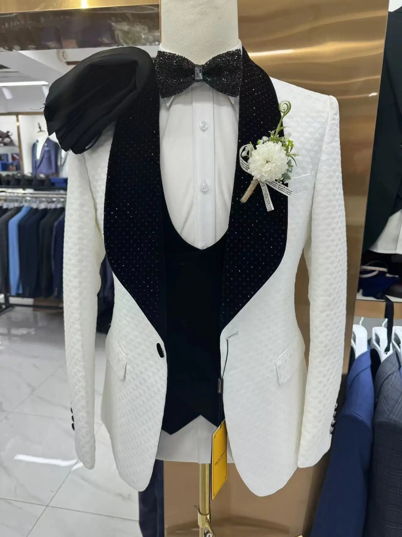 Elegant white tuxedo with black polka-dotted lapels displayed on a mannequin. Features a sparkling black bow tie, white shirt, and a floral boutonniere.