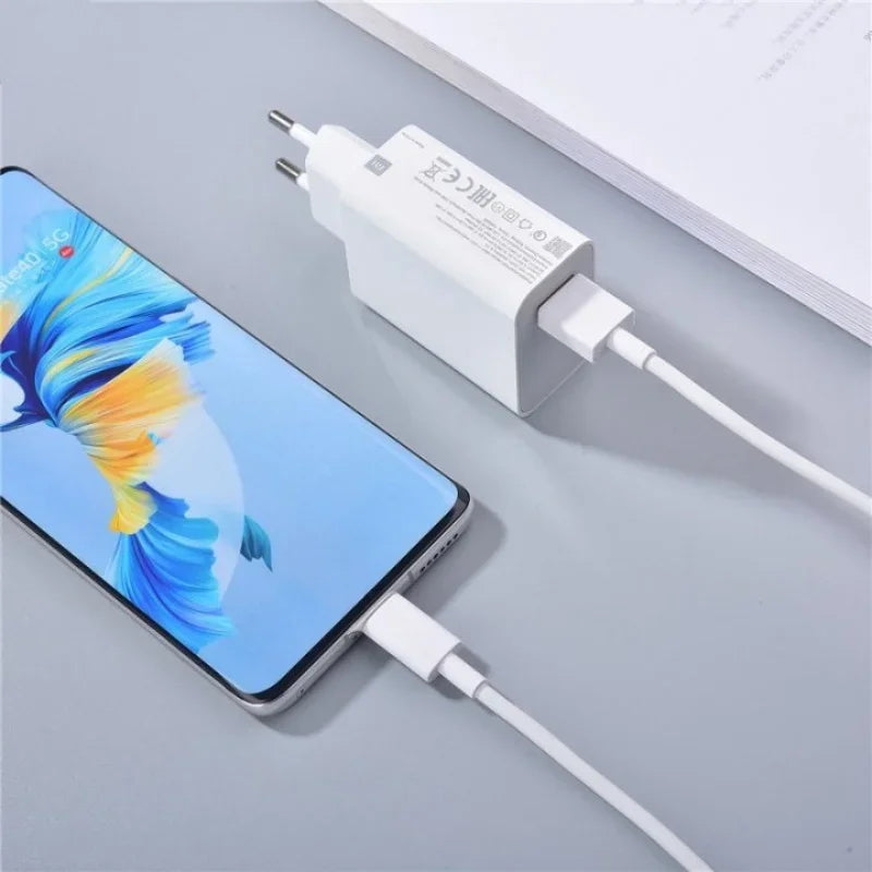 A smartphone with a colorful fish display is connected to a white rectangular charger via a USB cable, resting on a light gray surface near some documents.