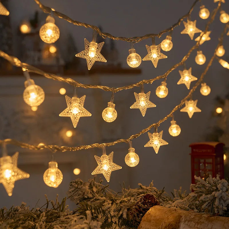 Warm white string lights featuring star and round bulb shapes create a cozy, festive atmosphere. The lights hang above snow-dusted greenery, hinting at a holiday setting.