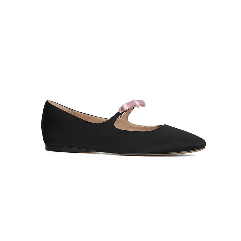 Black leather ballet flat with a pink satin bow strap. The shoe has a simple, elegant design, rounded toe, and flat sole, set against a plain white background.