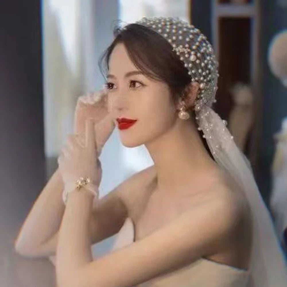 A woman in a strapless dress, wearing a pearl-decorated veil and earrings, with bright red lipstick, adjusts her gloves while seated in a softly lit room.