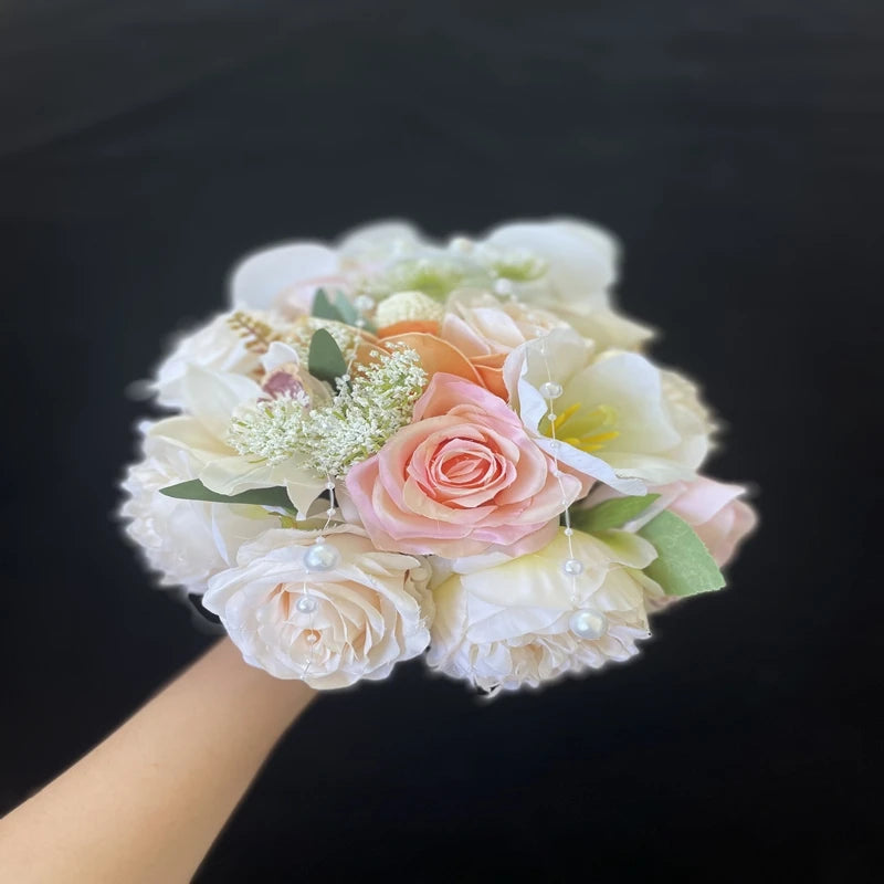 A hand holds a bouquet with pink and white roses, baby's breath, and green leaves, adorned with small white pearls, set against a dark background.