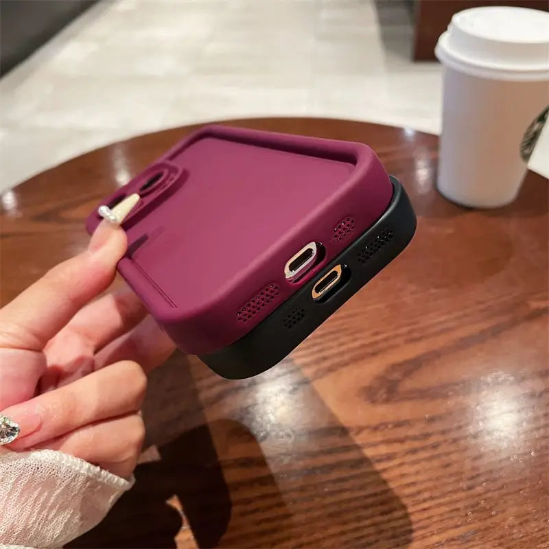 A hand holding a smartphone with a purple, thick case on a wooden table. A white disposable coffee cup is in the background.