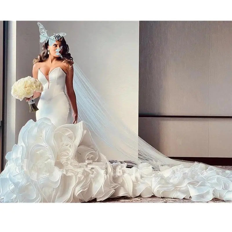 Bride in a white gown with a ruffled train and butterfly headpiece holds a bouquet of white roses, standing near a neutral-colored wall, looking to the side.