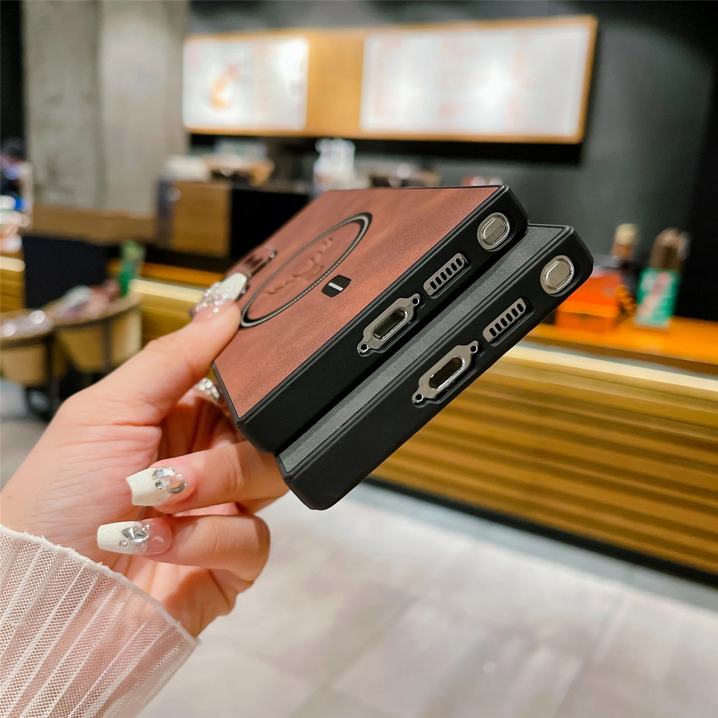A person holding two portable hard drives with wooden finishes, in a cafe setting. The drives have metal connectors, and the person has nails with white polish and silver designs.