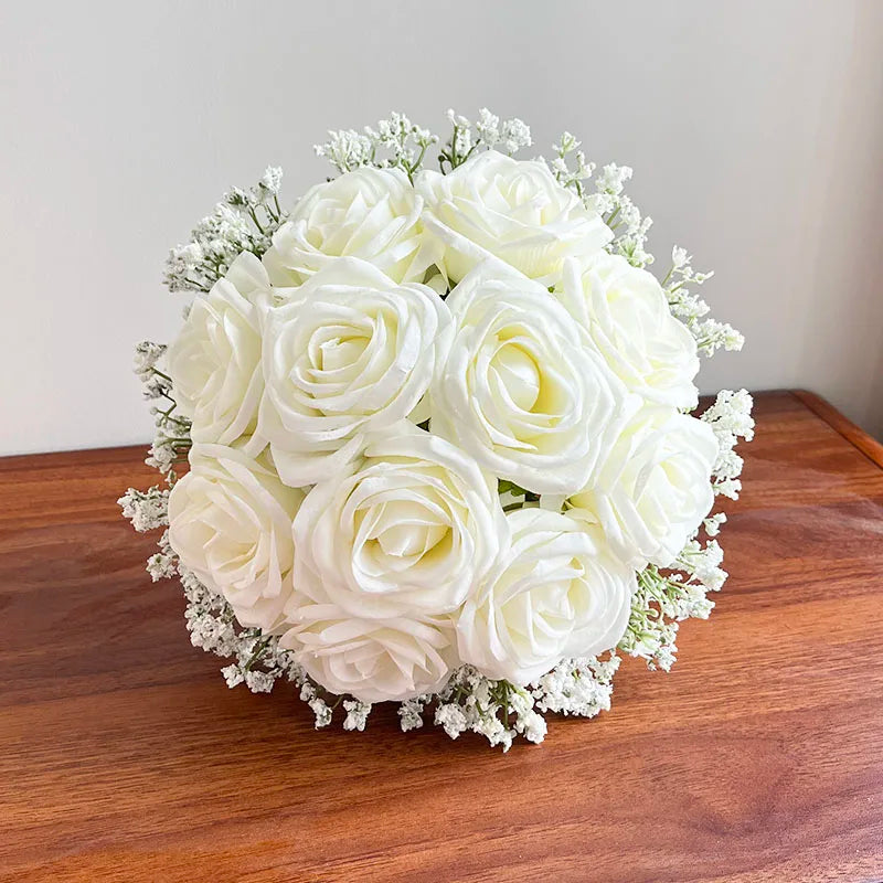 A wooden table showcases a bouquet of white roses surrounded by small white flowers, creating an elegant and delicate arrangement against a plain background.