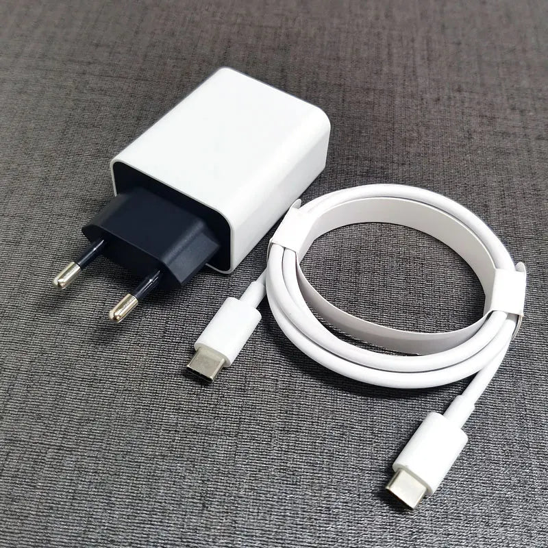 A white USB-C charger plug with two metal prongs sits next to a coiled white USB-C cable on a gray fabric surface.