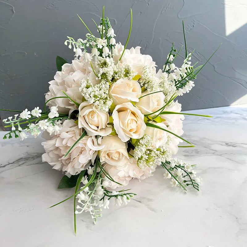 A bouquet featuring cream roses, white hydrangeas, and small white flowers, arranged with green sprigs, set on a marble surface against a textured gray background.