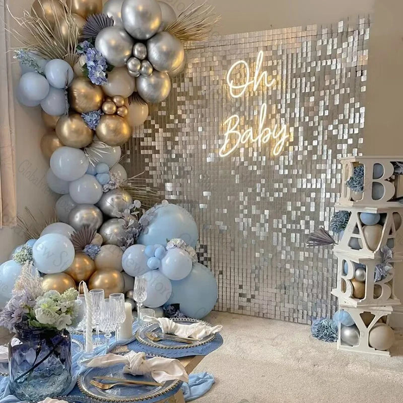 Baby shower setup with silver and blue balloons, a sequined backdrop with "Oh Baby" in neon, a decorated table with blue accents, and stacked letter blocks spelling "BABY."