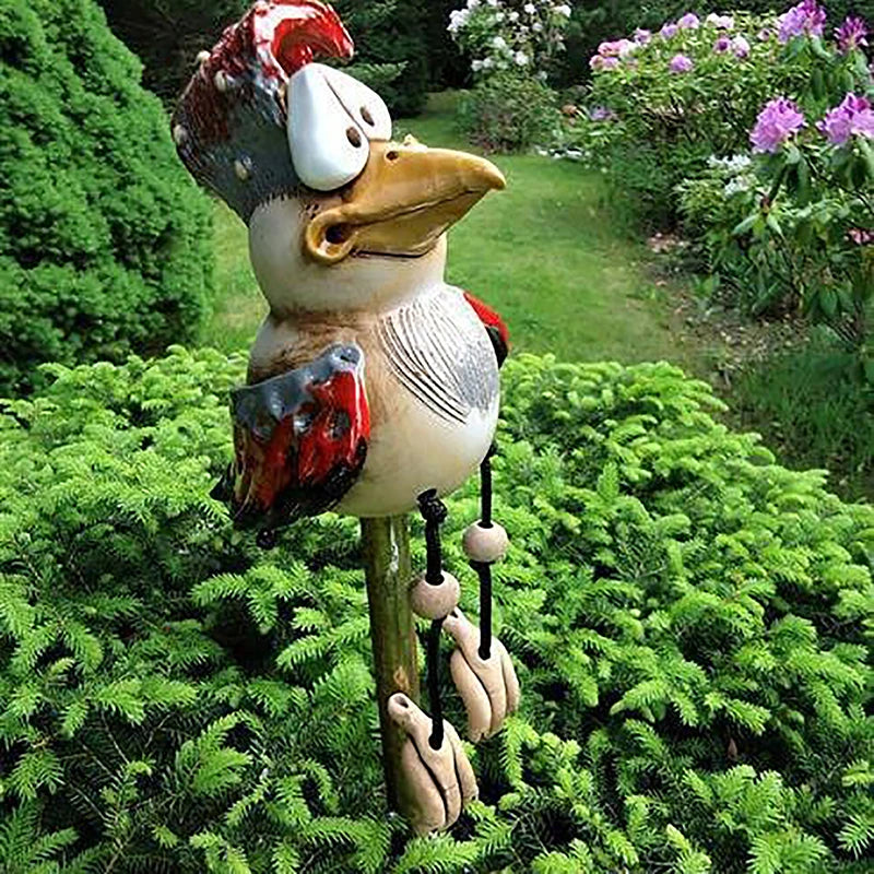 Whimsical bird sculpture with wide eyes and colorful wings perches on a lush green bush in a garden, surrounded by colorful flowers and a manicured lawn in the background.