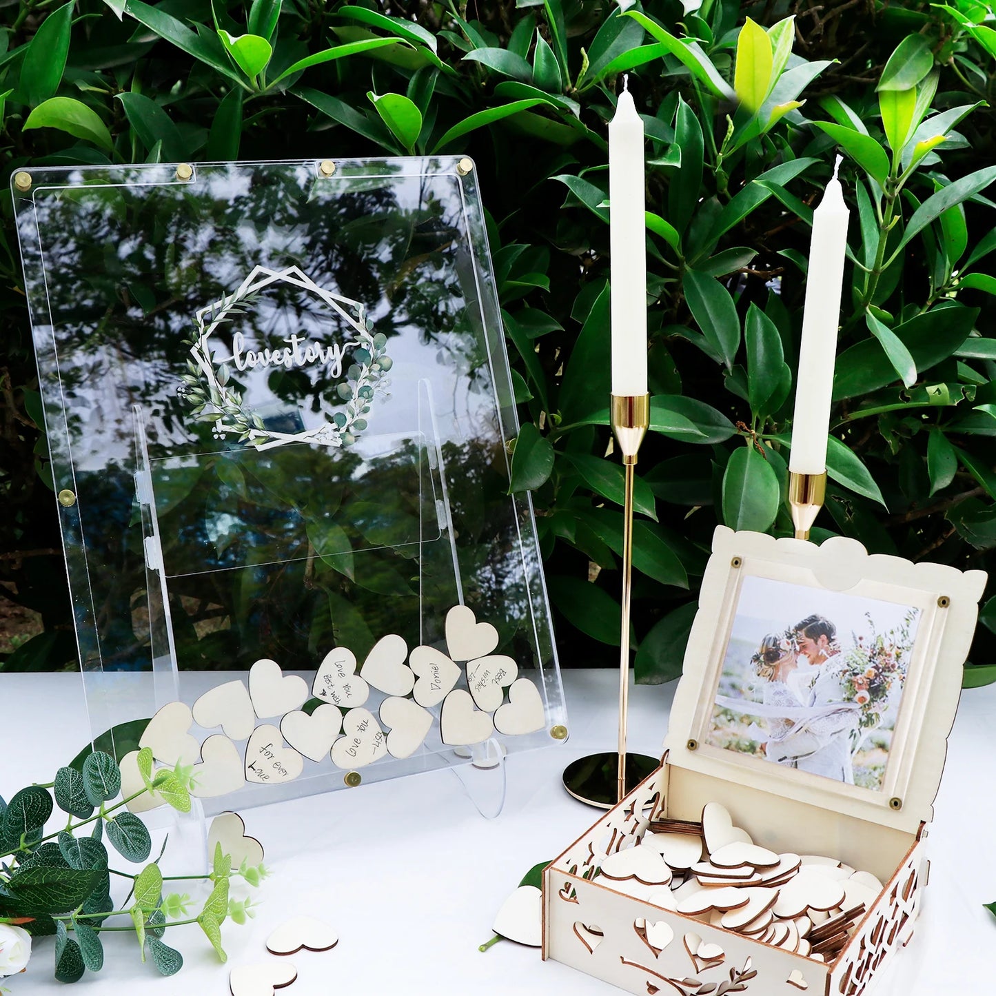 Elegant wedding display with acrylic sign reading "love story" and wooden heart tokens. Framed couple photo, two white candles with gold holders, and greenery in the background.
