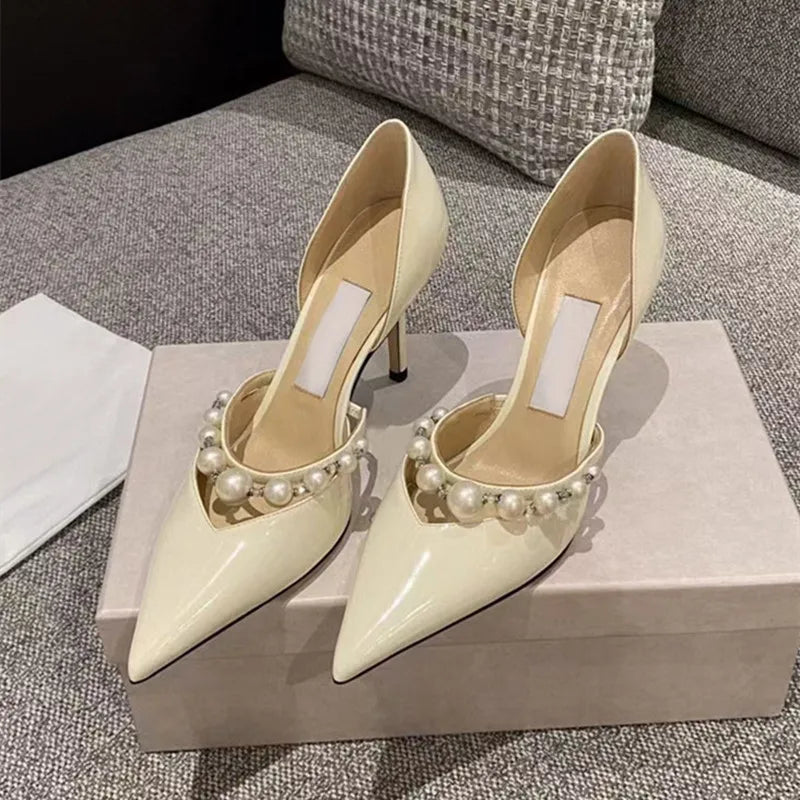 Cream-colored patent leather high heels with pearl accents sit elegantly atop a gray shoebox, placed on a textured gray sofa with a patterned cushion in the background.