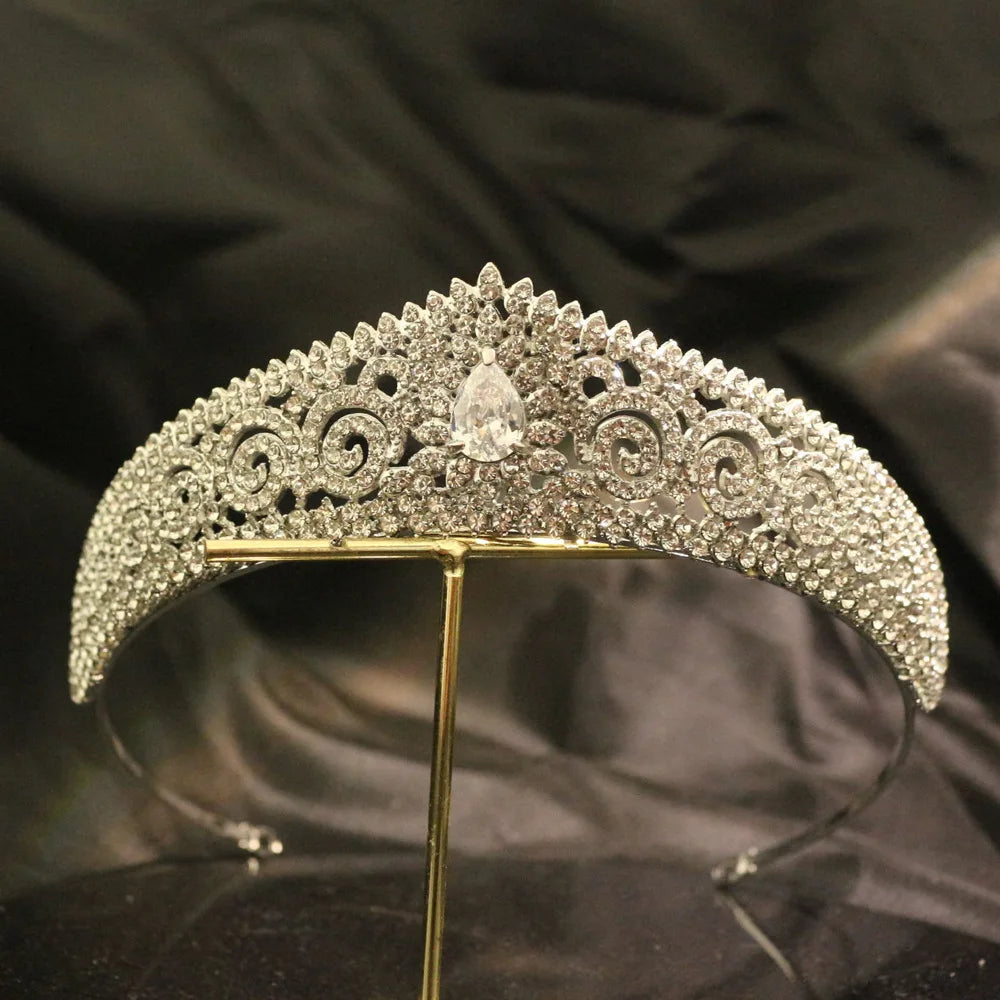 Elegant silver tiara adorned with intricate scrollwork and encrusted with numerous sparkling gemstones, centered with a pear-shaped gem, displayed against a dark background.