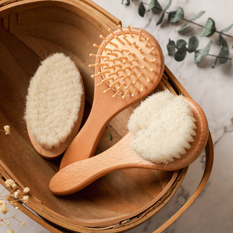 Wooden basket with three wooden bath brushes, featuring soft bristles and wooden pins, placed on a marble surface. Eucalyptus leaves and dried flowers surround the basket.