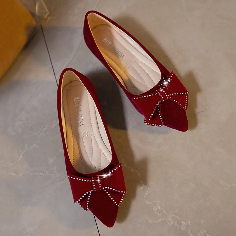 Red velvet flats with pointed toes, adorned with sparkling rhinestone-studded bows, are placed on a light-colored tiled floor.