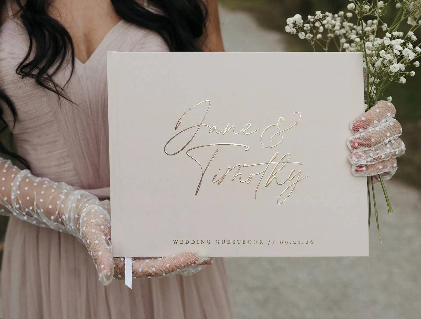 Woman in a blush dress and polka-dot gloves holds a wedding guestbook with "Jane & Timothy" in gold script, dated 09.21.26, alongside baby's breath flowers.