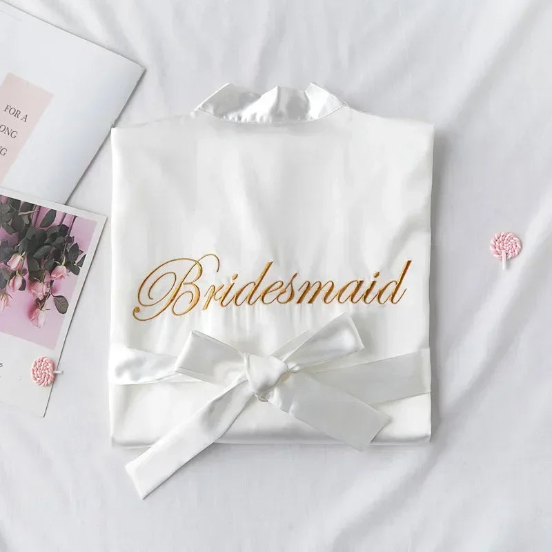 White satin robe with "Bridesmaid" embroidered in gold, tied with a ribbon. Placed on a bed near a floral-themed magazine and pink swirled candies.