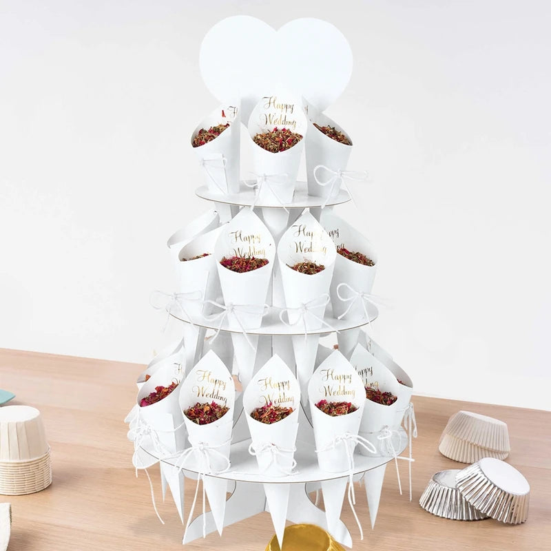 A tiered stand displays white cones filled with dried petals on a wooden table. Each cone reads "Happy Wedding" in gold script. A heart shape tops the arrangement.