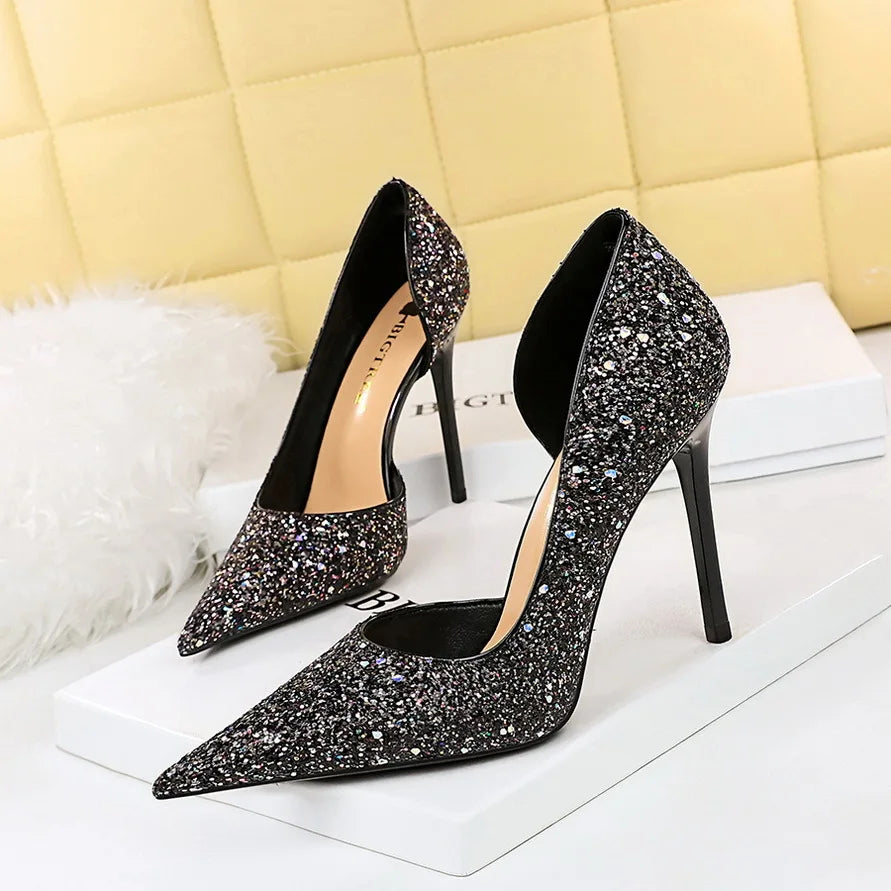 Black glittery high heels with pointed toes and stiletto heels are displayed on a white surface, against a yellow quilted background and a white fluffy accent.