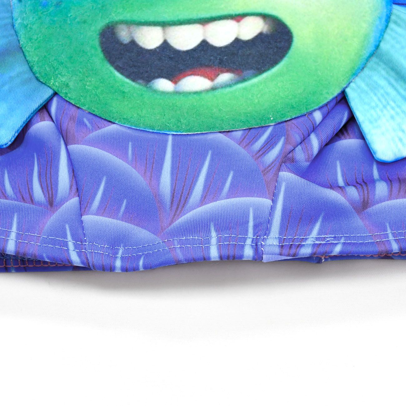 Close-up of a garment featuring a smiling green cartoon face with big teeth, on a purple patterned background with blue accents and stitched seams.