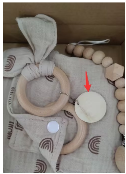 Wooden teething rings and beads on a beige patterned cloth with a bow. There's also a circular tag attached to the rings with a red arrow pointing at it.