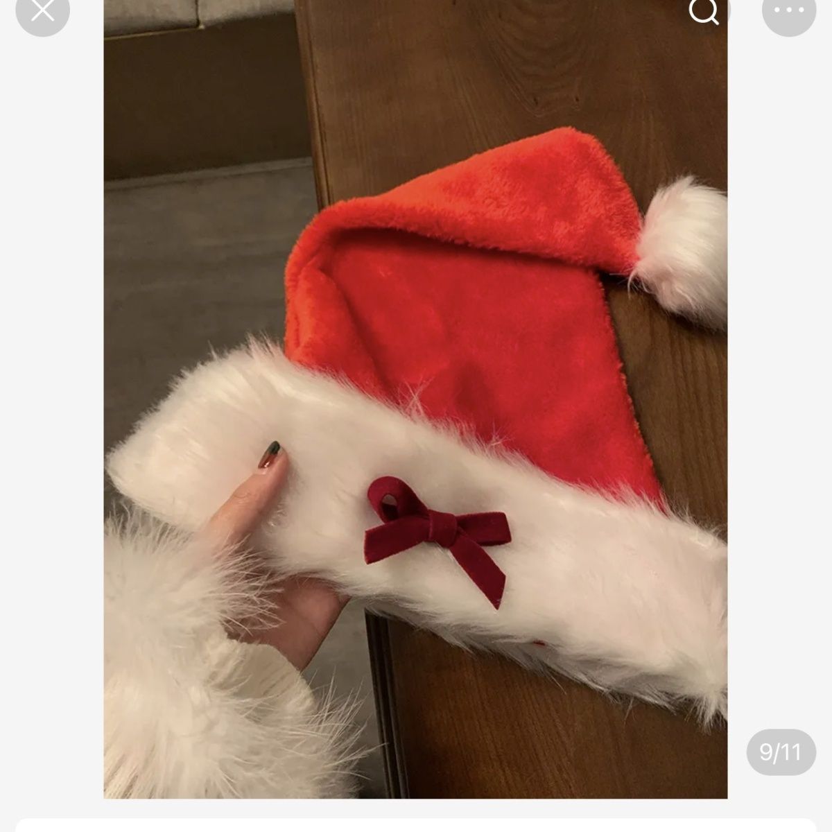 A hand holds a red Santa hat with white faux fur trim and a red bow, placed on a wooden table. The person wears a white fur cuff, suggesting a festive outfit.