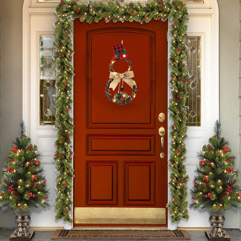 Festive front door with a snowman wreath and plaid hat. Doorway adorned with greenery, lights, and red berries. Two decorated small trees flank the entrance.
