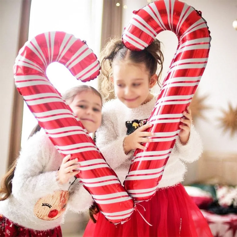 Two young girls in festive outfits hold large inflatable candy canes, smiling. One wears a sequin reindeer sweater, the other a red skirt, in a bright room with holiday decor.