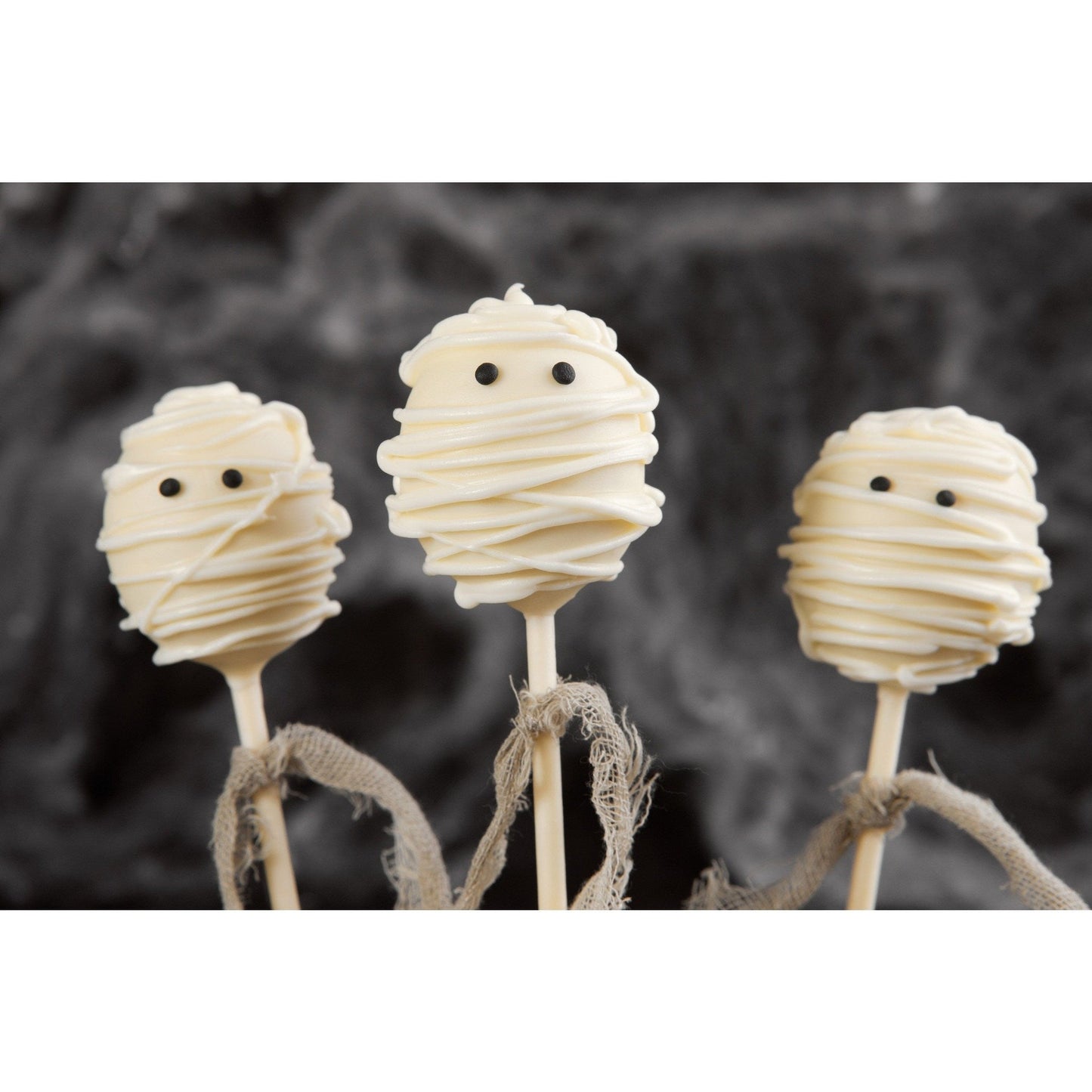 Three mummy cake pops wrapped in white icing with black eyes are arranged in front of a dark, blurred background. Each has frayed brown ribbon tied around the stick.