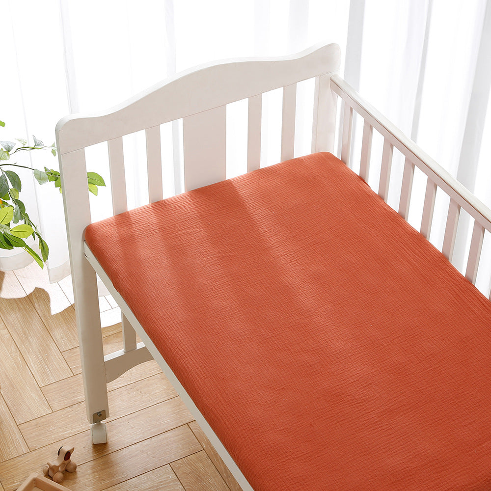 White crib with an orange mattress beside a window with sheer curtains, allowing natural light. A small plant and wooden toy are visible on the wooden floor.