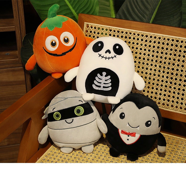 Four Halloween-themed plush toys sit on a chair: a smiling pumpkin, a skeleton with a ribcage design, a mummy with a green eye band, and a vampire in a tuxedo.