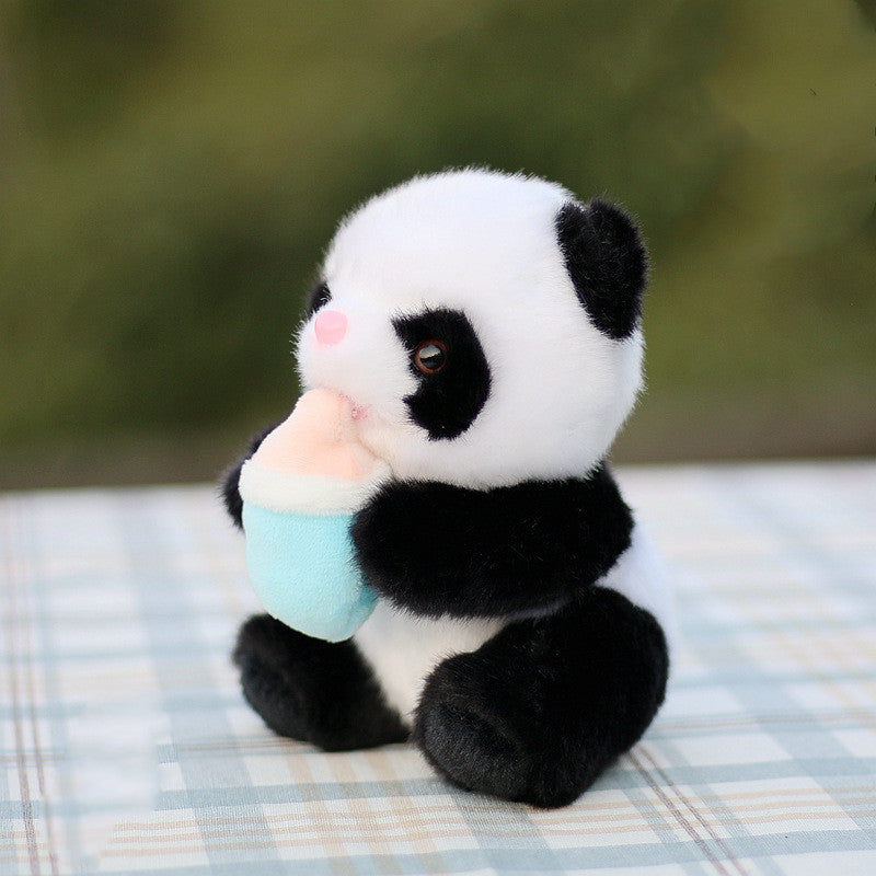 A plush panda toy holding a pastel blue bottle sits on a checkered surface. Its soft, black and white fur contrasts with the blurred greenery in the background.