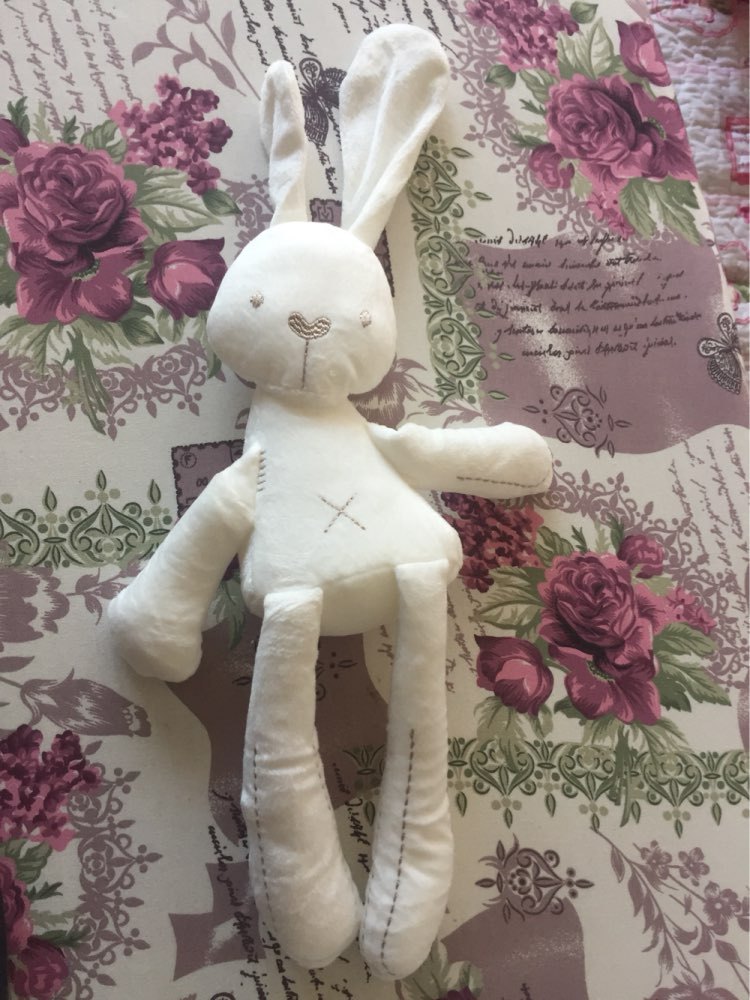 Plush white rabbit with stitched details lies on a floral and script-patterned fabric featuring pink and red roses with green leaves.