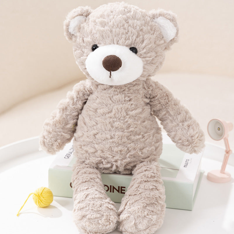 A soft, beige teddy bear with a white snout and brown nose sits on a book. Nearby, a small yellow ball of yarn and a pink lamp are visible on the table.