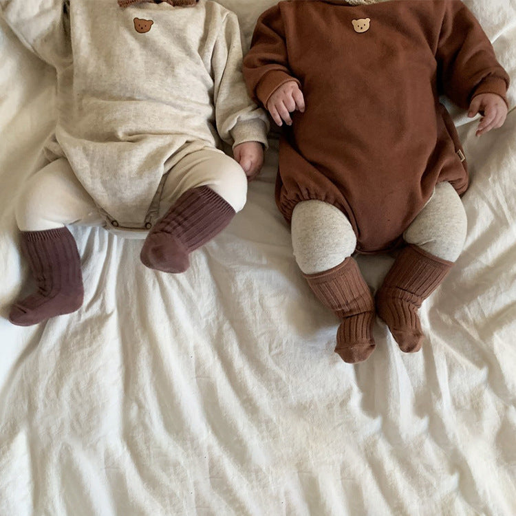 Two babies are lying on a white bed, wearing matching brown and beige outfits with bear motifs, and brown knee-high socks. They are positioned side by side.