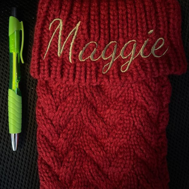 Red knit scarf with "Maggie" embroidered at the top, featuring a textured pattern. A green pen with a clip is placed beside the scarf on a dark surface.
