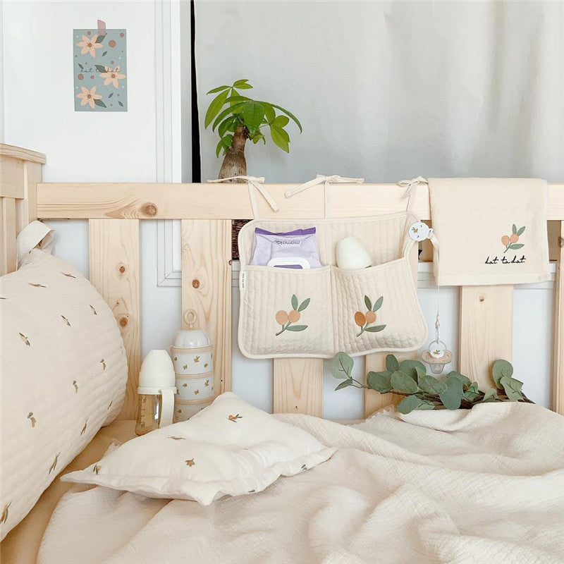 Cozy baby crib with beige bedding featuring small leaf patterns. A wooden rail supports hanging pouches filled with essentials, surrounded by a plant and a soft toy.