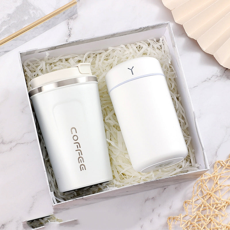 White coffee tumbler and insulated bottle in a gift box with shredded paper, displayed on a marble surface with a fan and netting nearby.