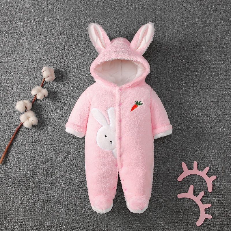 Pink baby onesie with bunny ears, a rabbit face, and carrot embroidery, laid on a gray surface. Cotton branch to the left and pink eyelash cutouts to the right.
