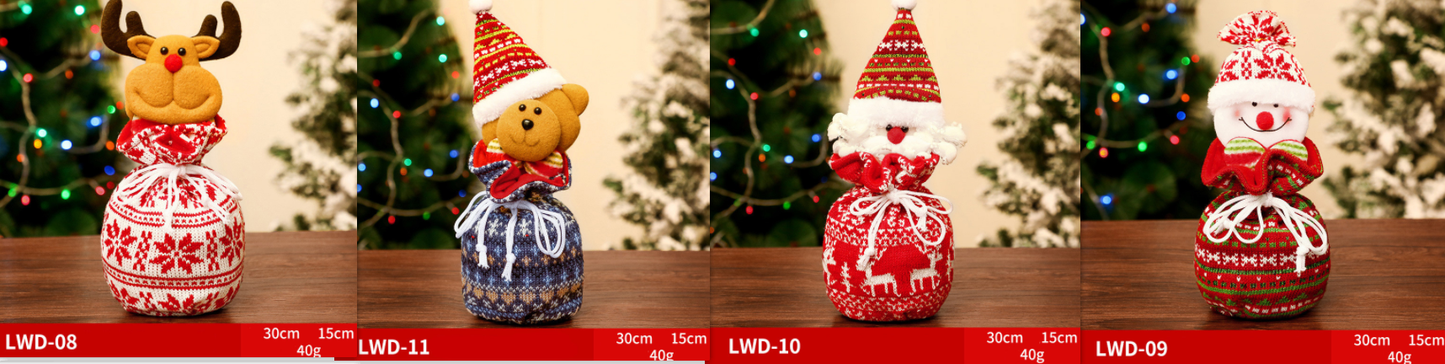 Four festive fabric gift bags with cute character tops: a reindeer, teddy bear, Santa, and snowman. Each bag is decorated in holiday patterns and set against a tree backdrop.