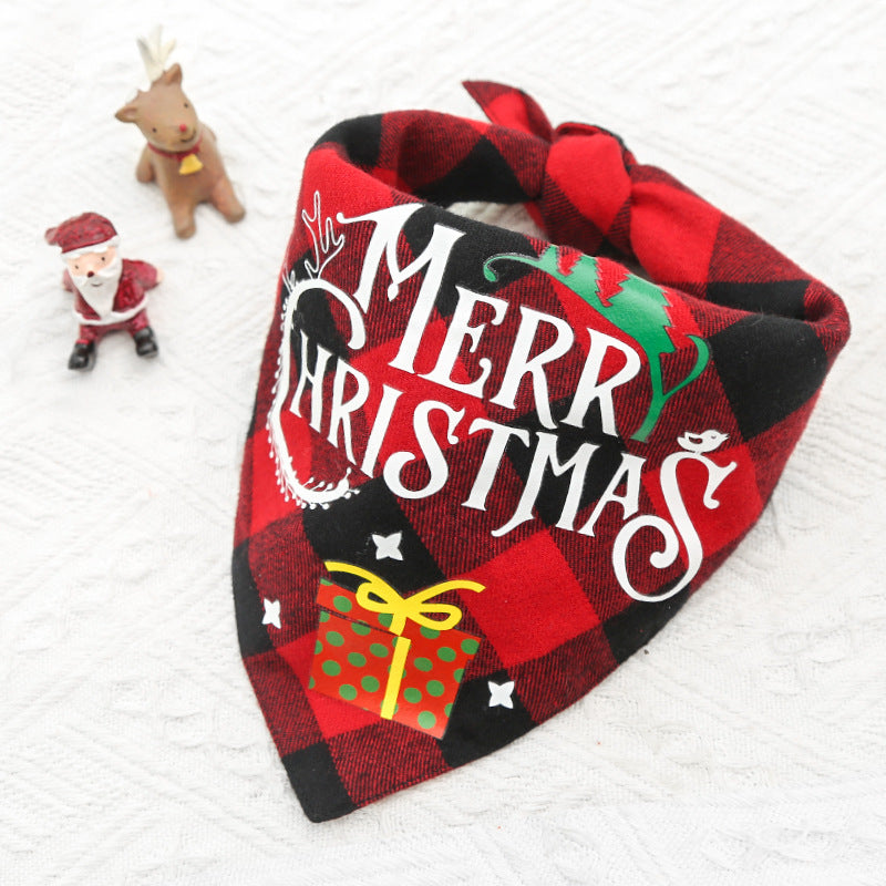 Red and black plaid dog bandana with "Merry Christmas" text, adorned with a small gift illustration. Nearby, a tiny Santa and reindeer figurine rest on a white textured surface.