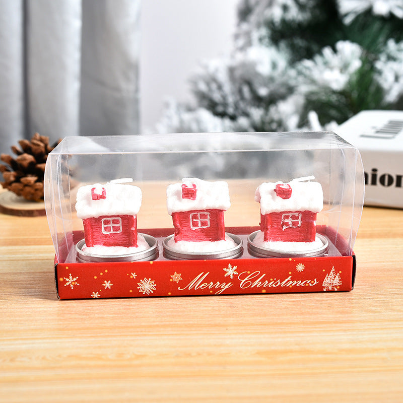 Three red house-shaped candles with snow-like roofs in a clear box on a wooden table. The packaging is red with snowflakes and reads "Merry Christmas." Pine cones in the background.