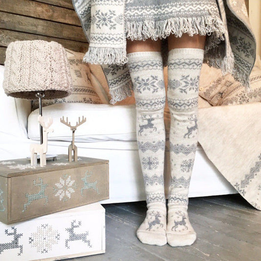 Cozy winter scene with a person wearing knitted snowflake and reindeer patterned over-the-knee socks and matching blanket. A reindeer-themed lamp and box sit beside them.