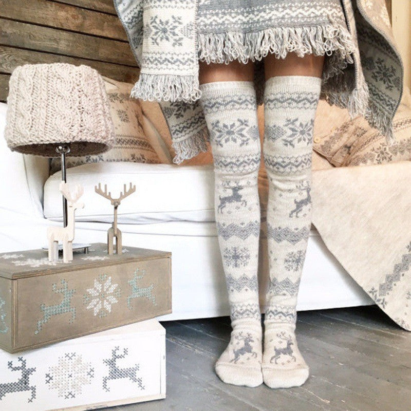 Cozy winter scene with legs in patterned knit socks and a matching fringed blanket. A wooden lamp and reindeer decor adorn a rustic box beside a white sofa.