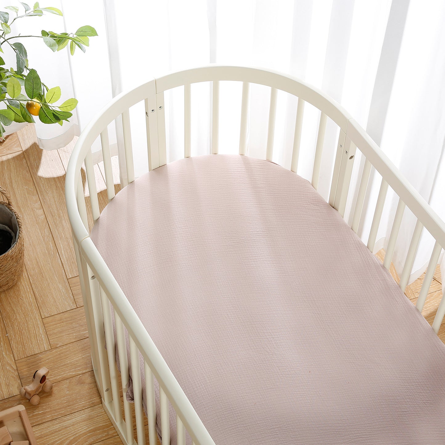 White oval crib with a pale pink sheet, set on wooden flooring. A wooden toy, potted plant, and soft natural light through sheer curtains complete the nursery setting.