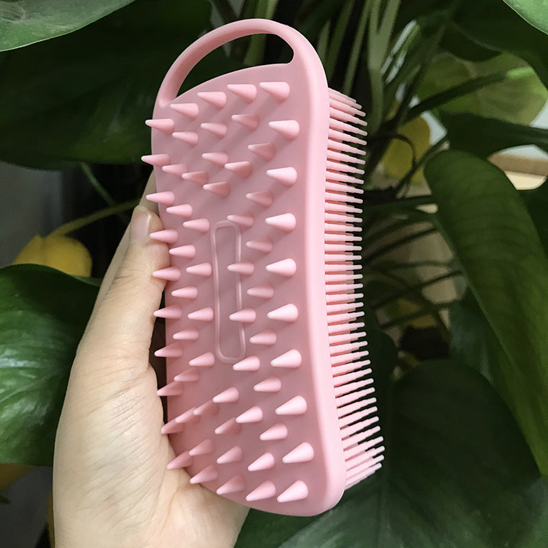 Hand holding a pink rubber scrub brush with rounded bristles on both sides, set against a backdrop of green leaves.