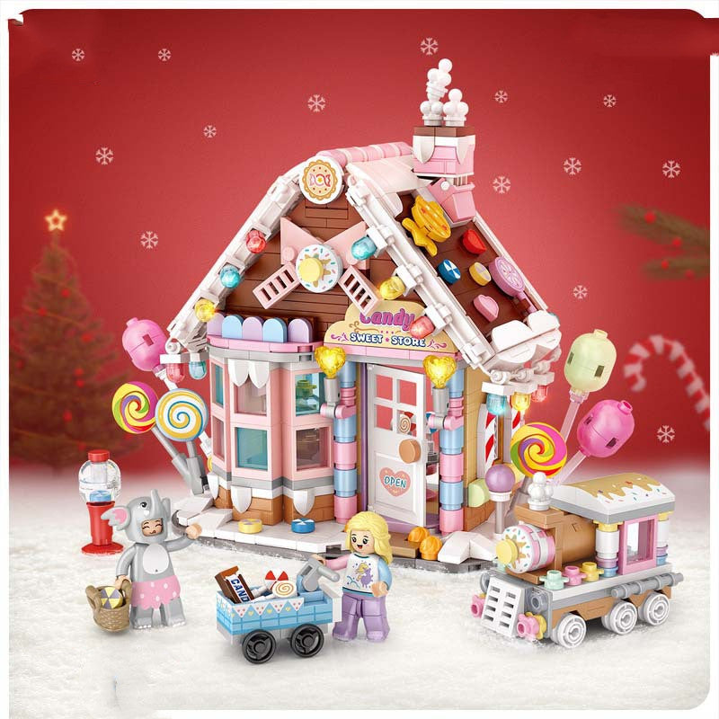 A festive toy set featuring a candy-themed house with colorful decoration, lollipop accents, a miniature train, and two toy figures. Snowflakes and a Christmas tree in the background.