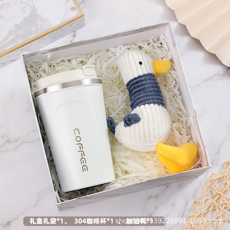 Gift box containing a white travel coffee mug with "COFFEE" text and a knitted toy duck. The items are nested in white shredded filler, placed on a marble surface.