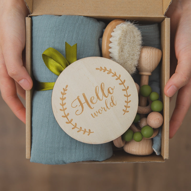 A hand holds a box containing a teal baby blanket with a green ribbon, a wooden brush, teether, and a round wooden plaque engraved with "Hello world" surrounded by a leafy design.